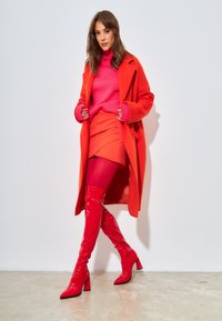 Abrigo de lana rojo sobre un suéter rosa, combinado con una falda roja y botas de muslo brillantes rojas. Texturas suaves y una combinación de colores audaz.