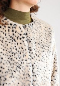 Faux-Fur-Jacke in cremigem Beige mit schwarzen Flecken, ausgestattet mit einer weichen Textur und einem runden Halsausschnitt, über einem grünen gerippten Rollkragenpullover getragen.