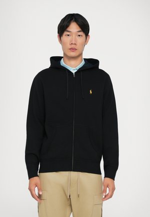 Haut à capuche zippé noir en tissu polaire, avec une poche avant, des cordons de serrage et un petit logo jaune sur le côté gauche de la poitrine.