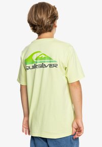 Camiseta de algodón de color amarillo claro con un gran logo en verde y negro de Quiksilver y un gráfico de montañas en la parte posterior, de manga corta y con un corte relajado.