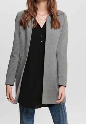 Femme portant des colliers en or superposés, une tunique noire boutonnée, un blazer gris ouvert sur le devant et un pantalon bleu texturé, debout devant un fond clair.