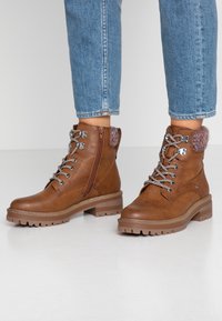 Bottes courtes en cuir brun avec une finition texturée, lacets à l'avant, fermeture éclair latérale et accent en tissu à motif sur le talon. Semelle en caoutchouc épaisse.
