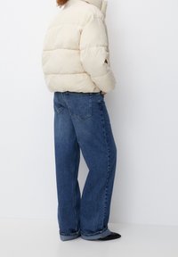 Giacca a vento beige con design trapuntato, abbinata a jeans in denim blu medio a vestibilità ampia. Entrambi i capi presentano una consistenza notevole e una silhouette rilassata.