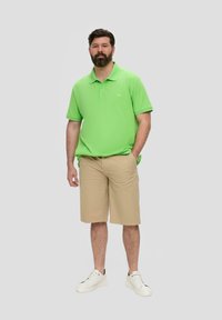T-shirt polo vert, shorts beiges, baskets blanches. Manches courtes, col classique et coupe décontractée. Un homme se tient avec les mains dans les poches.