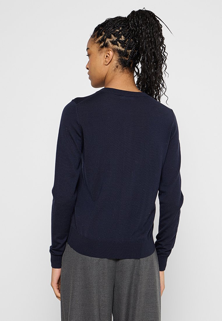 Frau mit geflochtenem Haar in einem Pferdeschwanz, die einen marineblauen Langarmpullover und graue, lockere Hosen trägt, mit dem Rücken zu einer einfachen Hintergrund. 