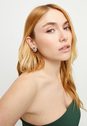 Pendientes de plata con un diseño de ojo verde y acentos circulares en capas, sobre un fondo claro. La modelo tiene el cabello largo y ondulado.