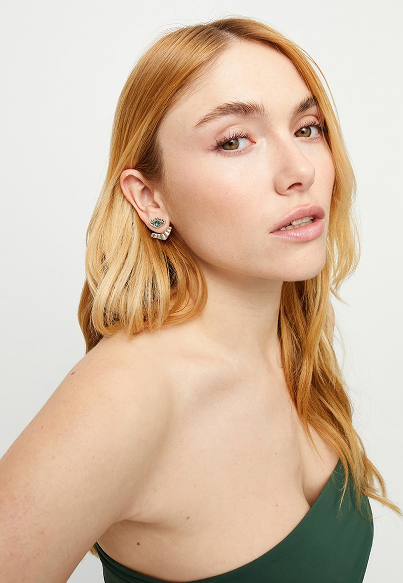 Pendientes de plata con un diseño de ojo verde y acentos circulares en capas, sobre un fondo claro. La modelo tiene el cabello largo y ondulado.