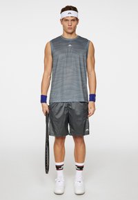Abbigliamento sportivo con una maglietta grigia senza maniche con righe ondulate, pantaloni corti grigi abbinati e scarpe da ginnastica bianche con accenti neri. Tiene una racchetta da tennis.