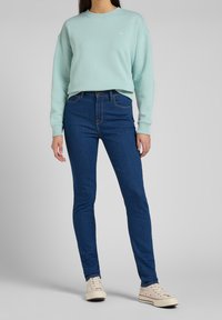 Persona con felpa azzurra chiara, jeans skinny blu scuro e sneakers bianche a bassa punta, in piedi contro uno sfondo chiaro e neutro.