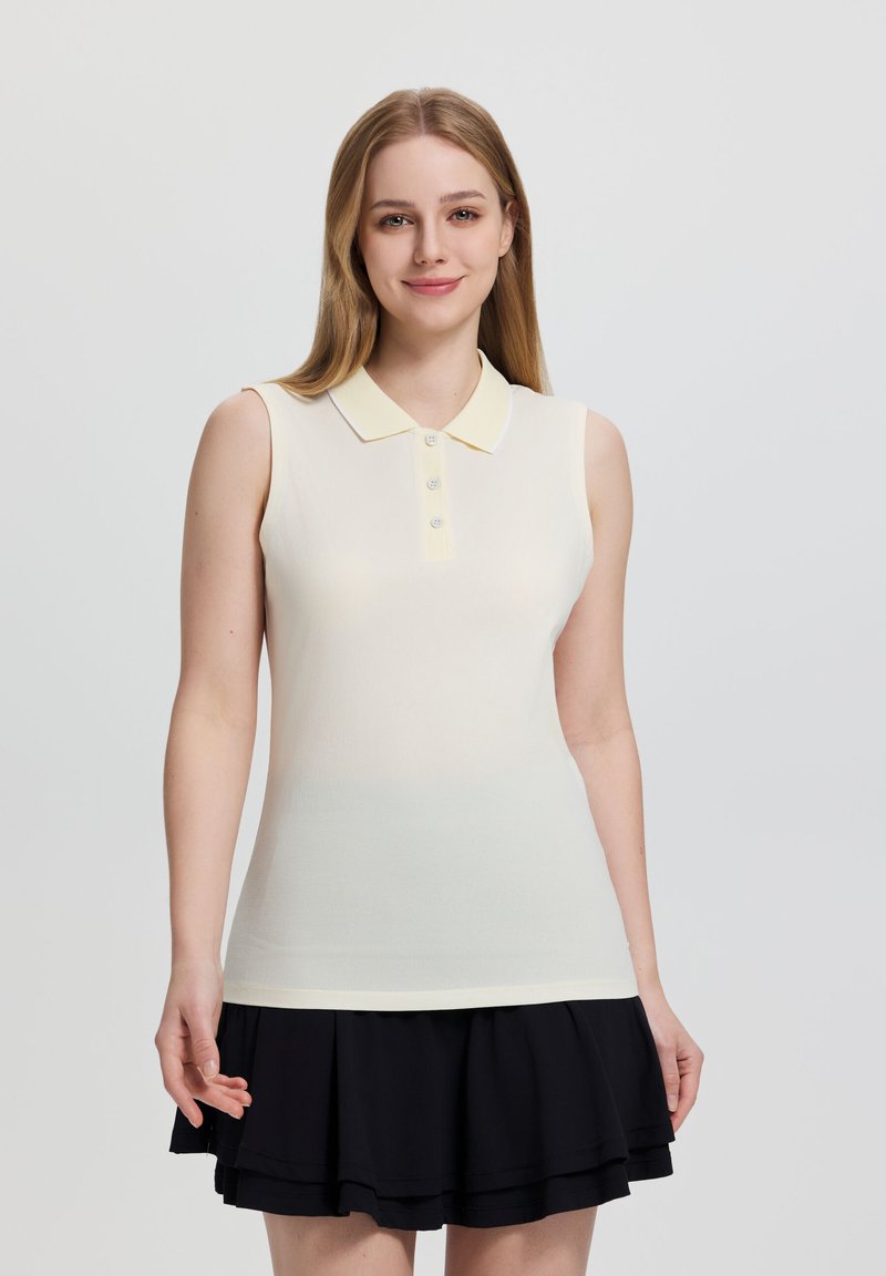 Giovane donna con lunghi capelli biondi che indossa una polo giallo pallido senza maniche e una gonna nera a strati, sorridente e in piedi davanti a uno sfondo semplice.