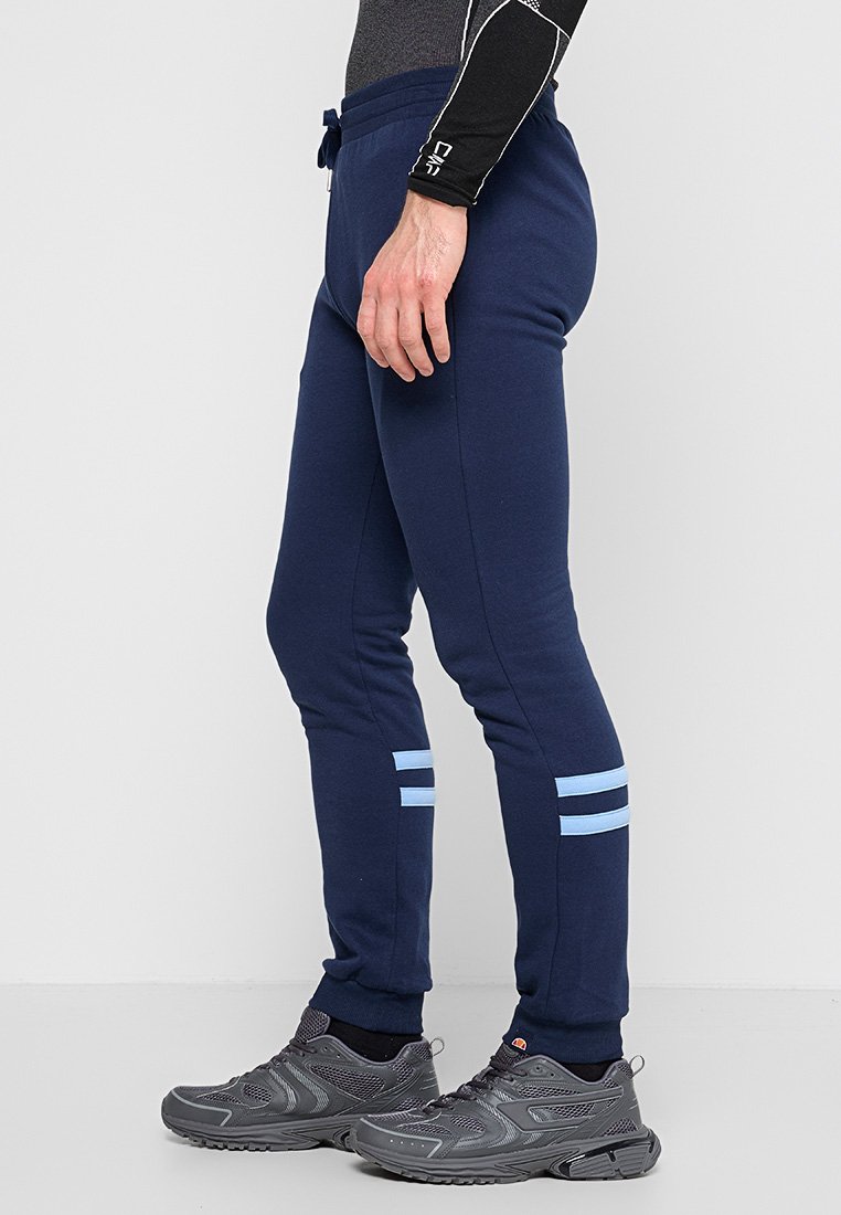 Mann in navyblauen Jogginghosen mit hellblauen Streifen an den Waden und dunkelgrauen Laufschuhen, seitlich stehend vor weißem Hintergrund.