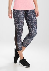 Czarno-szare legginsy w geometryczny wzór, z przezroczystymi panelami po bokach, połączone z czarnymi butami sportowymi.