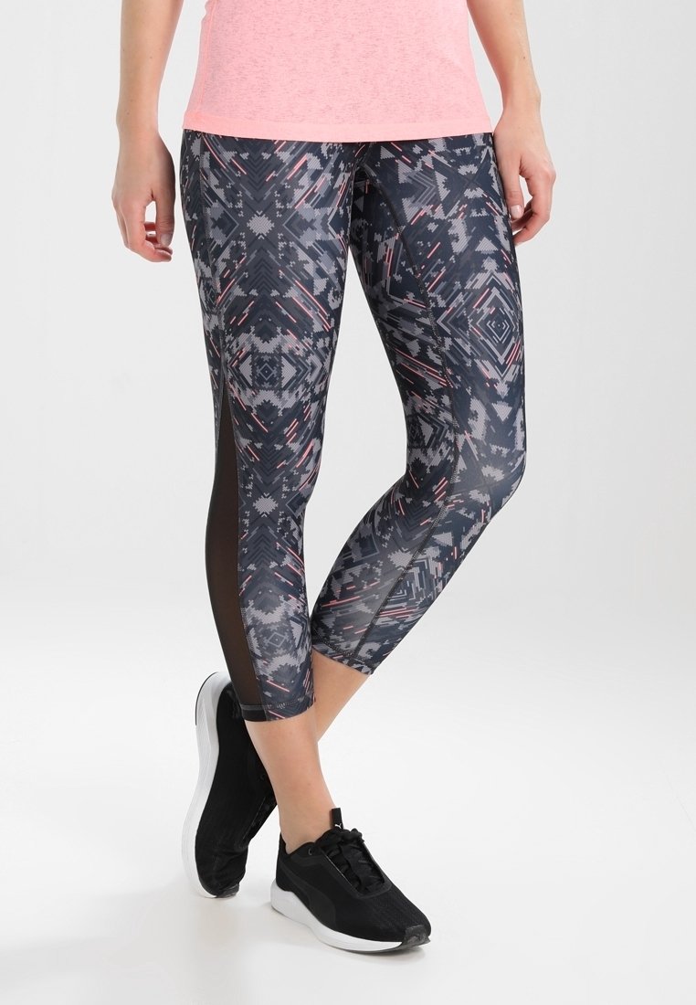 Czarno-szare legginsy w geometryczny wzór, z przezroczystymi panelami po bokach, połączone z czarnymi butami sportowymi.