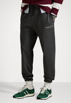 Mann trägt dunkelgraue Polo Ralph Lauren Jogginghose, weiße Socken und grüne Sneakers mit weißen Schnürsenkeln, Hände in den Taschen, vor einem einfarbigen Hintergrund.