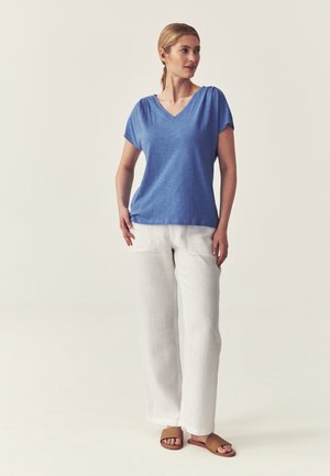 Vrouw staat, draagt een blauwe korte mouw V-hals top, witte losse broek en bruine sandalen met open tenen, kijkt naar rechts met handen bij de zakken.