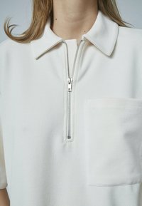 Hvid, ribbet polo skjorte med lynlås halvvejs og en brystlomme. Den har en struktureret krave og en glat tekstur.