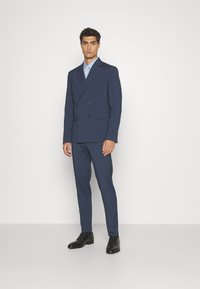 Marineblaues, doppelt geknöpftes Sakko mit Revers, kombiniert mit einer passenden Hose und schwarzen Lederschuhen. Strukturierter Stoff, taillierte Passform.