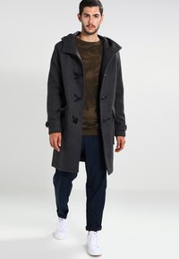 Cappotto duffle grigio scuro in lana con cappuccio, bottoni a bilanciere e tasche laterali, indossato sopra una camicia con motivo camouflage e pantaloni blu navy.