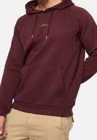 Sweat à capuche bordeaux avec une poche kangourou, des cordons de serrage ajustables et des poignets côtelés. Présente un petit logo sur la poitrine et une texture douce.