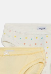 Dois pares de roupa interior infantil: um em creme com estampa de corações multicoloridos, outro em amarelo claro liso com um laço e acabado em rendas.