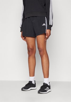 Person iført sorte Adidas-shorts og sorte sneakers med hvide sokker, stående mod en ensfarvet lys baggrund.