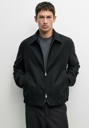 Giovane uomo con capelli corti che indossa una giacca nera con zip sopra una maglia grigia, in piedi con le mani in tasca davanti a uno sfondo semplice.