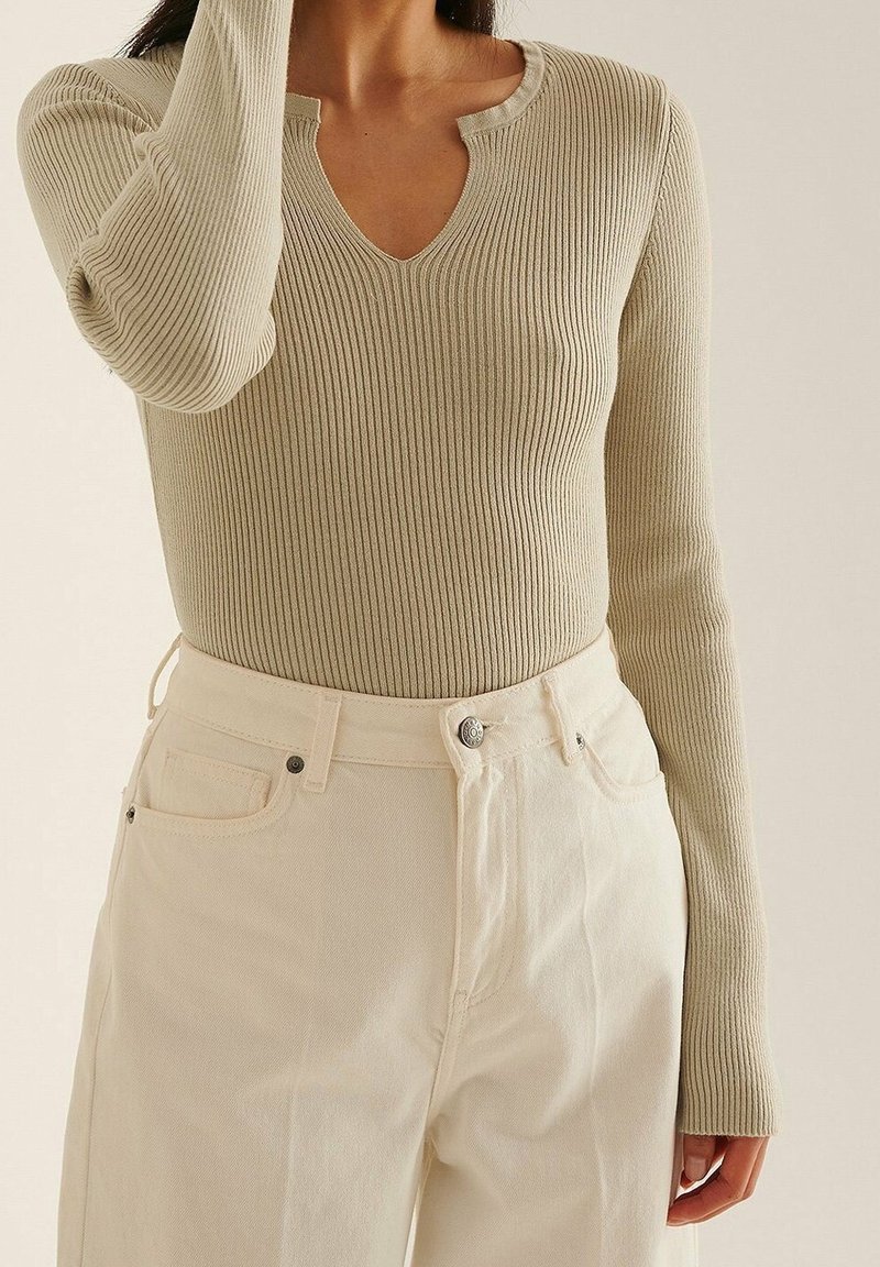 Haut beige à manches longues côtelé avec col en V, associé à un pantalon taille haute blanc cassé, porté par une personne debout avec un bras levé.