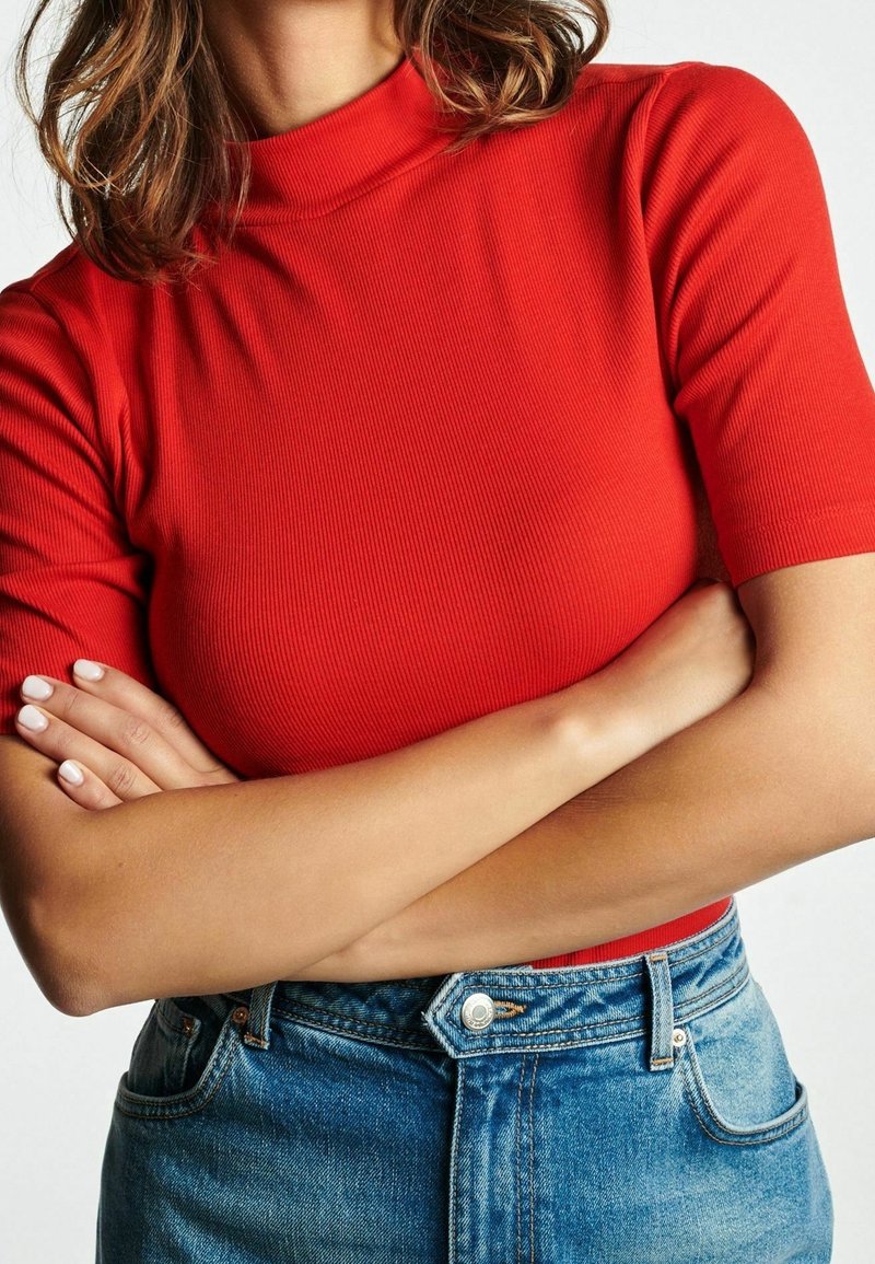 Personne les bras croisés portant un haut rouge à manches courtes et un jean en denim bleu, montré du cou à la taille.