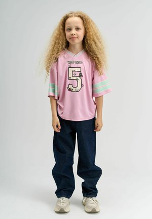 Roze sportshirt met bloemenpatroon en het nummer 5, gecombineerd met donkerblauwe baggy broek en neutraalgekleurde sneakers.
