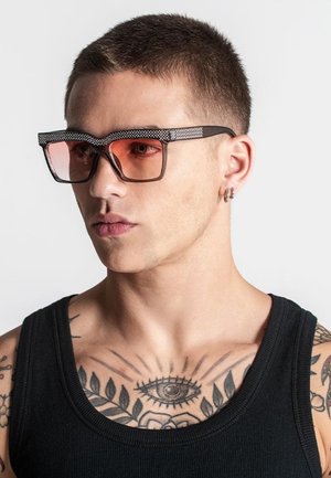 Sorte solbriller med orange tonet linser og teksturerede sorte arme. Båret af en person med tatoveringer, iført en sort tanktop.