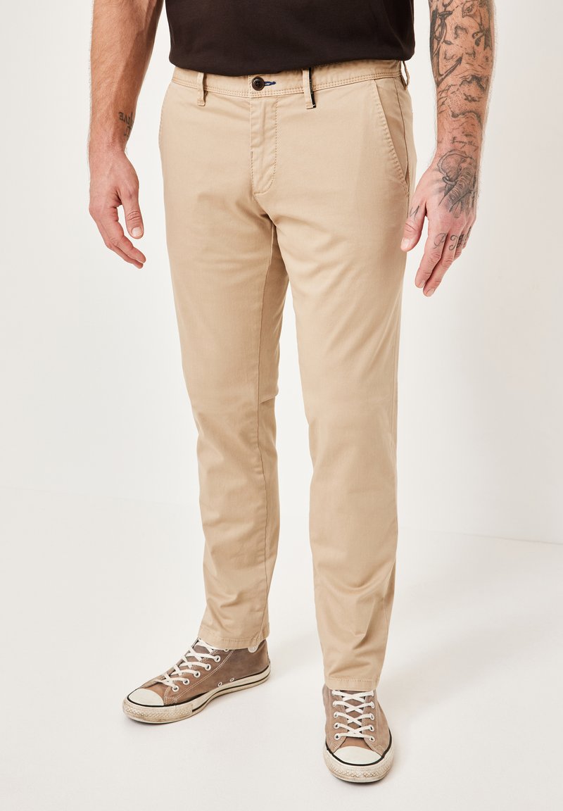 Beige Chinos mit schlanker Passform, ausgestattet mit Seitentaschen und einem einzelnen Knopfverschluss. Kombiniert mit braunen Segeltuch-Sneakern, geschnürt und getragen.