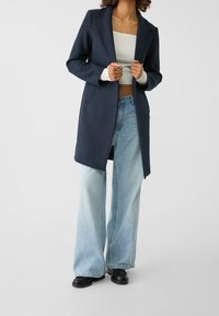 Marineblå lang frakke i uldblanding med skrå kraver, sidelommer og lige pasform, kombineret med lyseblå bukser med brede ben og sorte sko.