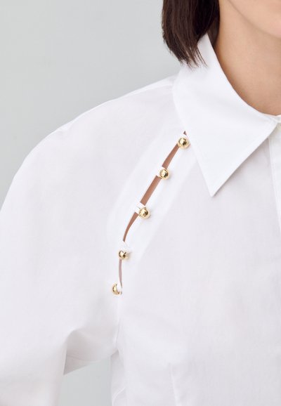 In primo piano, una camicia bianca con bottoni decorativi dorati lungo un'apertura laterale e un colletto classico, che mette in risalto elementi di design moderno.