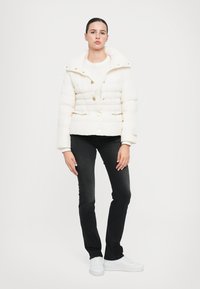 Giacca a vento in morbido tessuto bianco, con taglio cropped, grande colletto, bottoni dorati e tasche con zip; abbinata a jeans neri e scarpe bianche.