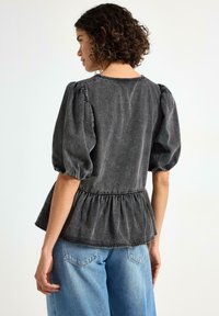 Kvinde med krøllet hår iført en sort top med puffærmer og samlet talje samt lyseblå denim jeans, der står foran en ensfarvet baggrund.