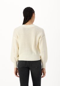 Crème-kleurige gebreide trui met een textuurpatroon, losse pasvorm en geribde zoom. Ballonmouwen lopen taps toe bij de manchetten. G gedragen met donkere jeans.