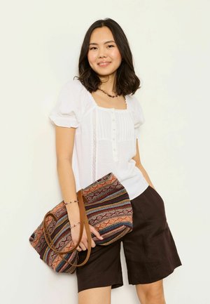 Jeune femme aux cheveux mi-longs portant un chemisier blanc et un short sombre, tenant un grand sac fourre-tout coloré à motifs.