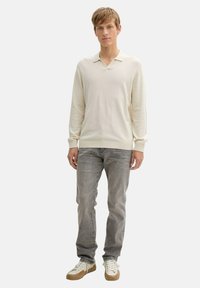 Világos bézs pulóver V-nyakkal és hosszú ujjal, szürke farmernadrággal és krémszínű, barna talpú sportcipővel párosítva. Egyszerű, textúrált kötött mintázat.