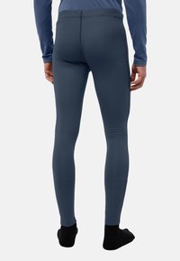 Uomo che indossa leggings termici aderenti blu scuro e una maglia a maniche lunghe abbinata, che sta in piedi a piedi nudi su uno sfondo bianco.