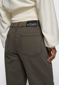 Nahaufnahme einer Person, die braune Hosen mit elastischem Bund und Gesäßtaschen trägt, versehen mit einem schwarzen "10 DAYS"-Label, kombiniert mit einem weißen Langarmshirt.