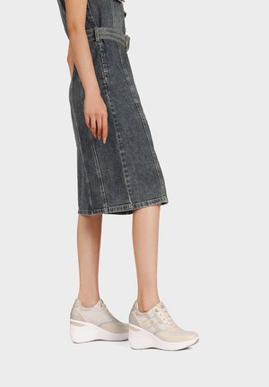 Donna che indossa una gonna in denim nero sbiadito di media lunghezza e sneakers beige con zeppa, in piedi di lato su uno sfondo chiaro uniforme.