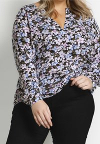 Frau in einer langärmligen blumigen Bluse mit blauen, lila und weißen Blumen, kombiniert mit schwarzen Hosen vor einem neutralen Hintergrund.
