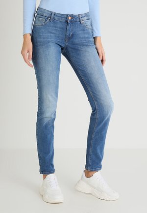 Slim-fit blauwe jeans gedragen met witte chunky sneakers en een lichtblauw topje met lange mouwen op een staand persoon tegen een effen achtergrond.