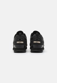 Svarta sportskor med slät syntetöverdel, dämpad häl, texturerad sula och metalliska logodetaljer bak.