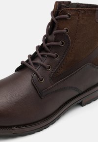 Botín de tobillo de cuero marrón y textil con punta redonda, sistema de cordones y texturas contrastantes; presenta ojales de metal y una suela de goma.