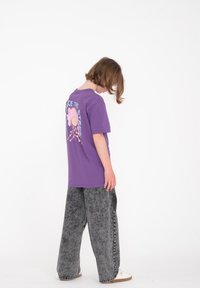 Camiseta de algodón morada con estampado gráfico en la parte posterior, combinada con pantalones anchos grises y zapatos blancos. Diseño sencillo con un ajuste relajado.