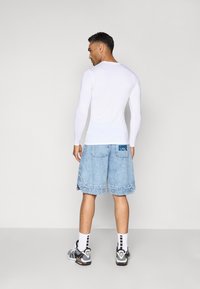 Mand, der vender væk, iført en hvid lang�ærmet skjorte, lyseblå denimshorts med et ryglogo, hvide sokker og sort-hvide sneakers.