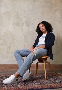 Blazer navy, camiseta gráfica blanca, pantalones vaqueros desgastados de un azul claro y zapatillas blancas con acentos negros, sentado en una silla de madera.
