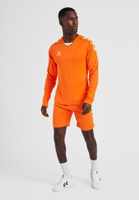 Oranje sportshirt met lange mouwen, bijpassende shorts en witte accenten. Bevat logo op de borst en een gestructureerde stof. Draagt het met witte sneakers.