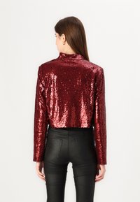 Saspiests sarkans sequinēts tops ar augstu apkakli un garām piedurknēm, apvienots ar melniem pieguļošiem biksēm. Tekstūra ir spīdīga un atstarojoša.
